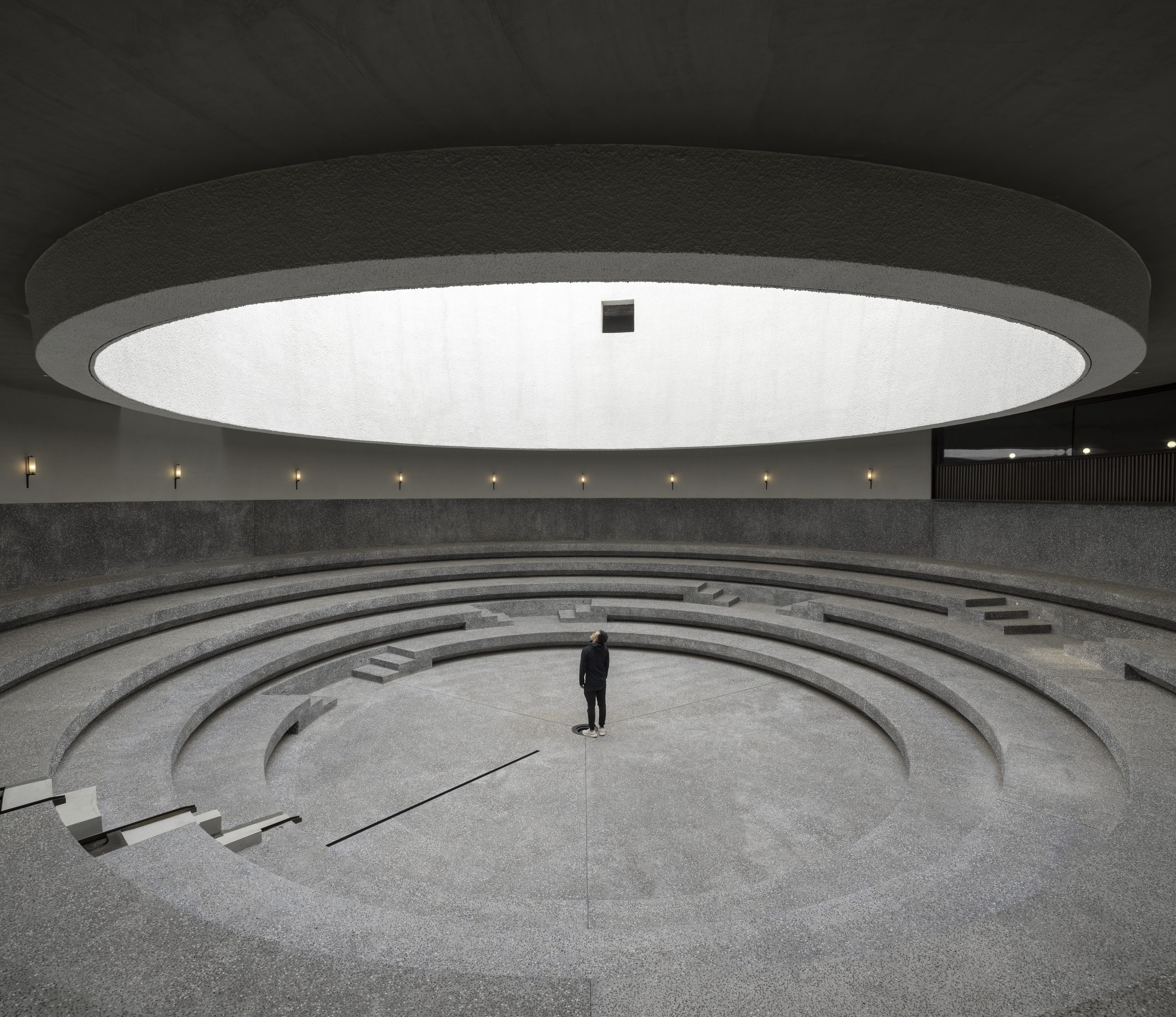 A person stands alone in the center of a large, circular, amphitheater-like space with concentric concrete steps and a round skylight above.