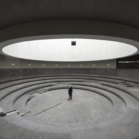 A person stands alone in the center of a large, circular, amphitheater-like space with concentric concrete steps and a round skylight above.