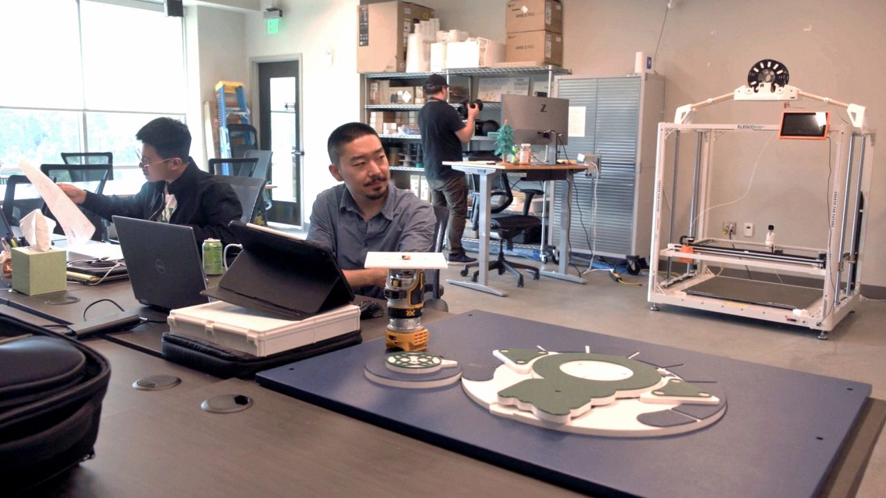 Three people work in a modern lab with electronics, laptops, a large 3D printer, and mechanical parts on the table.