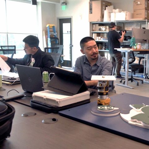 Three people work in a modern lab with electronics, laptops, a large 3D printer, and mechanical parts on the table.