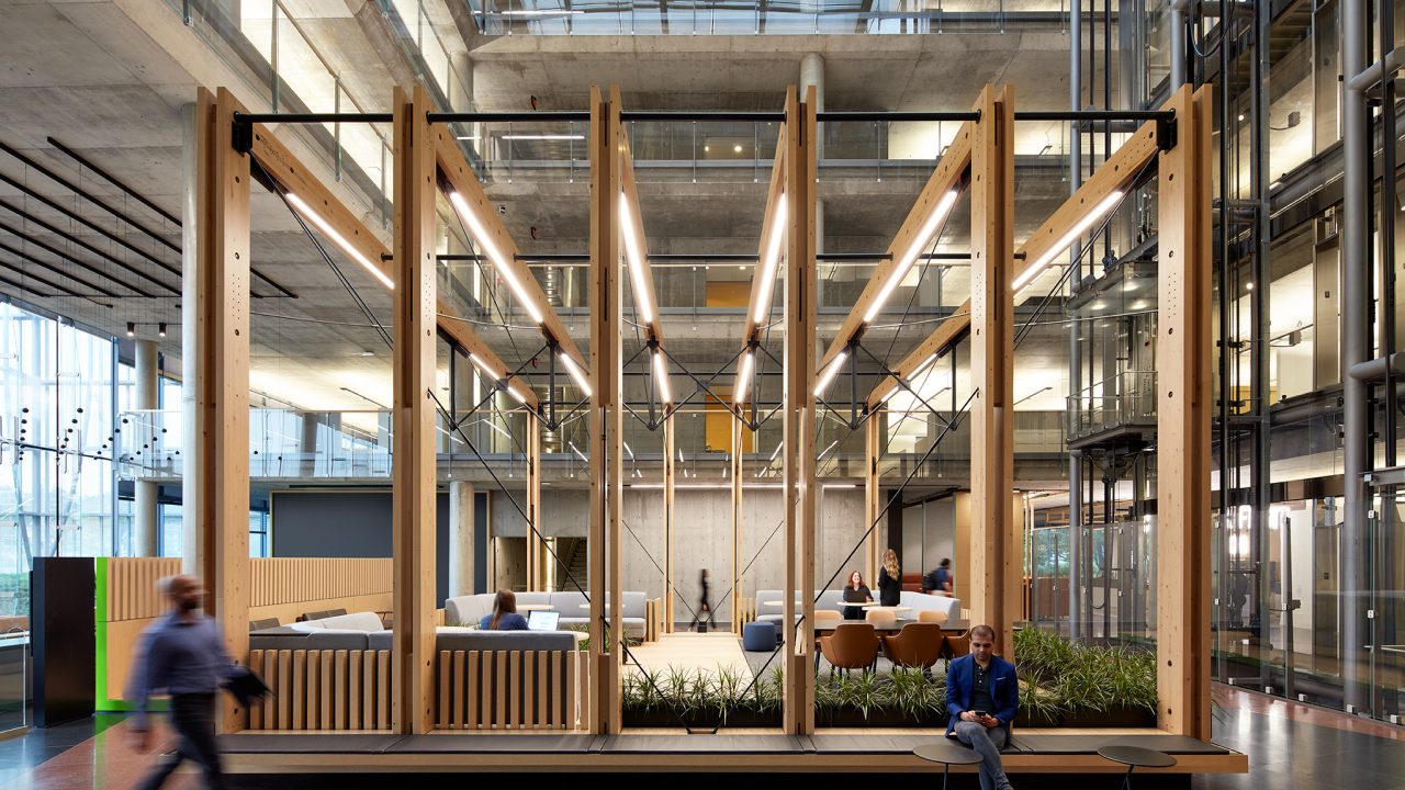 Modern office space with exposed concrete, glass walls, wooden beams, and people sitting or walking in a communal seating area.