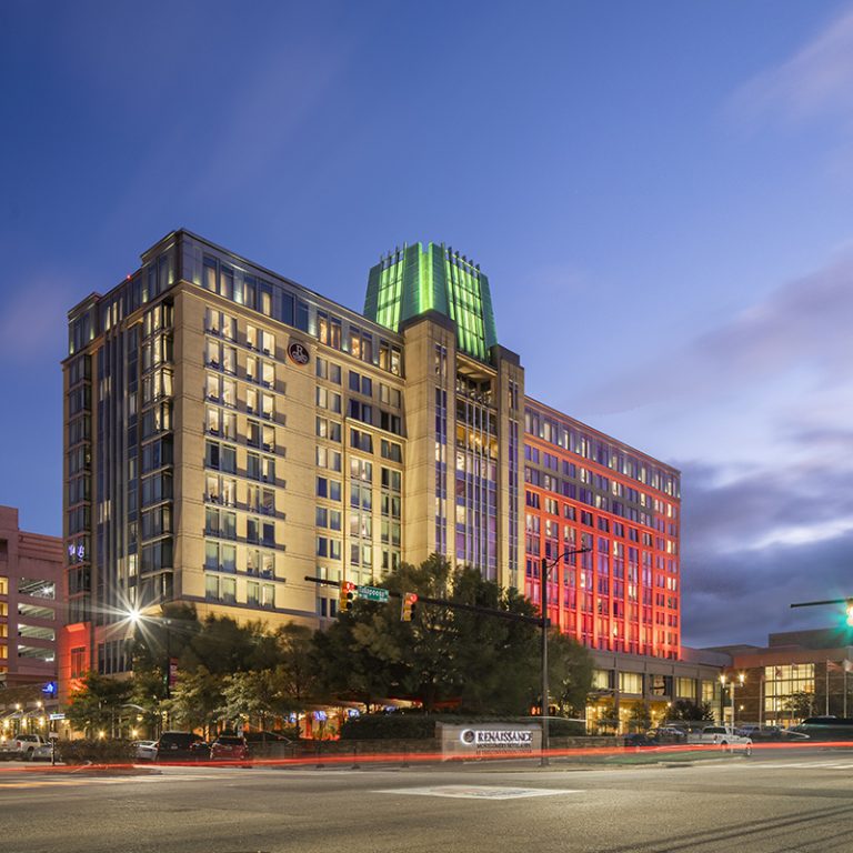 A multi-story hotel building illuminated at night with a green-lit dome on the roof, colorful exterior lighting, and traffic lights at a city intersection in the foreground.