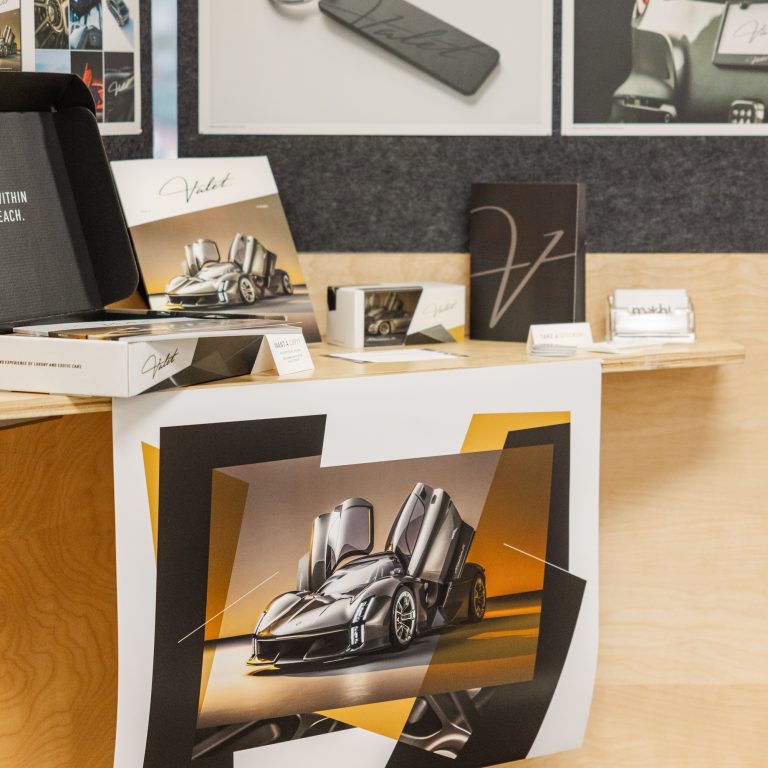 A display of automotive-themed products including sketches, a keychain, notebooks, business cards, and a large poster featuring a sports car with open butterfly doors.