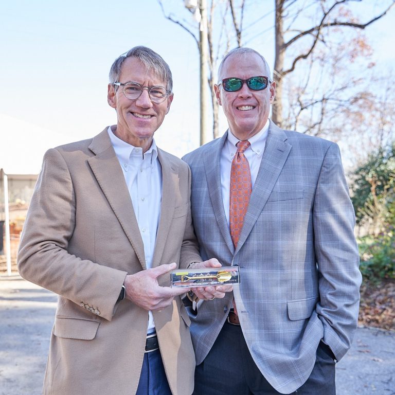 Two men in suits stand outdoors, smiling at the camera. The man on the left holds a rectangular glass award or plaque. Leafless trees and a clear sky are in the background.