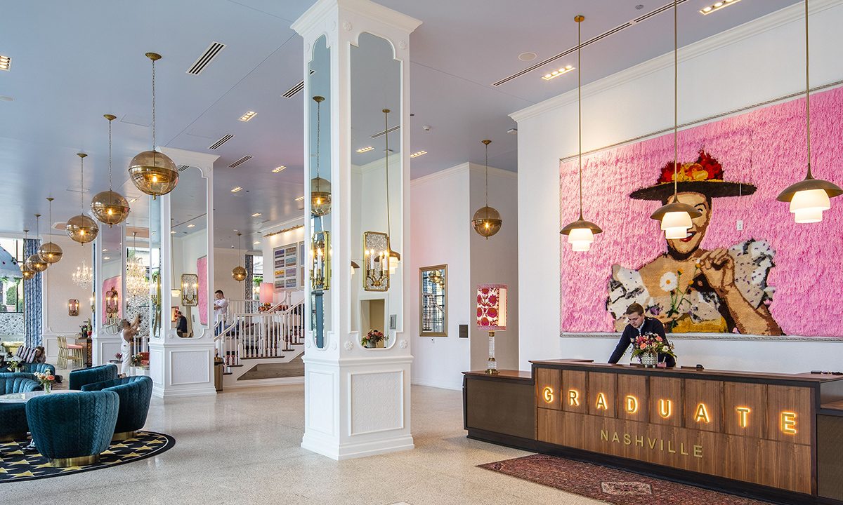 Hotel lobby with modern decor, teal seating, round gold pendant lights, a front desk labeled Graduate Nashville, and a large bright pink portrait of a woman on the wall.