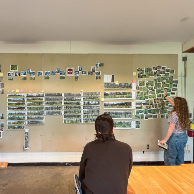 Two people review and arrange landscape photographs and notes on a large bulletin board in a room with a wooden table.