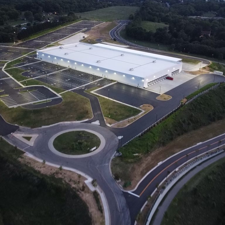 Aerial view of a large, white industrial building with surrounding parking lots, access roads, a roundabout, and green landscape at dusk.