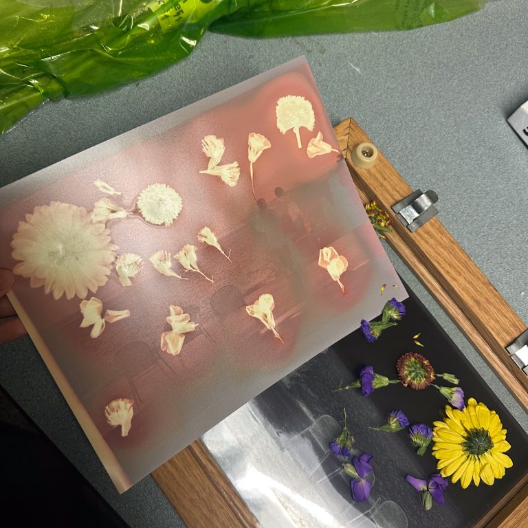 A hand holds a sheet showing white flower imprints next to a wooden board with fresh yellow and purple flowers arranged on it. Green floral wrapping sits nearby.