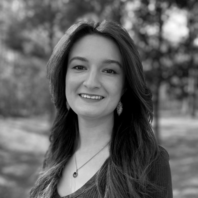 A woman with long dark hair smiles at the camera while standing outdoors, with trees and blurred greenery in the background.
