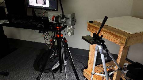 A camera on a tripod is positioned in front of a computer monitor and a wooden table with a white surface in a room.