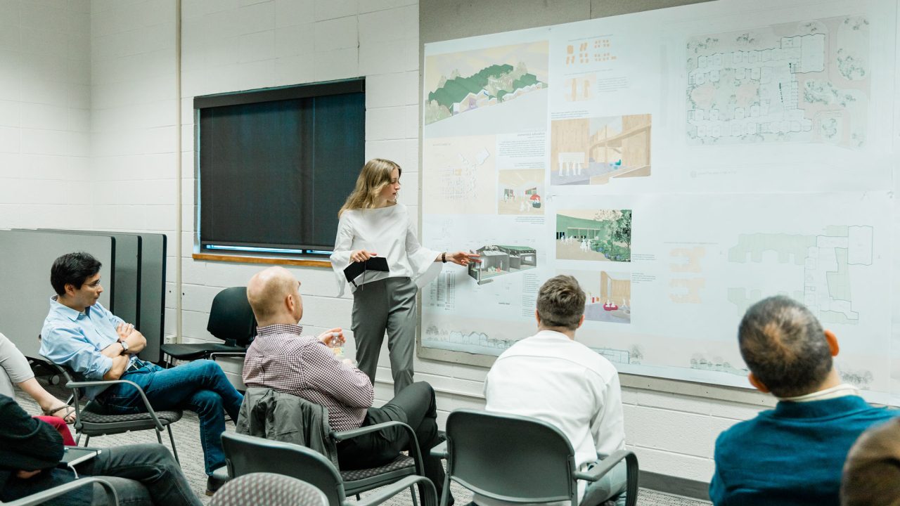 A woman stands and presents architectural plans on a wall to a seated group in a conference room.