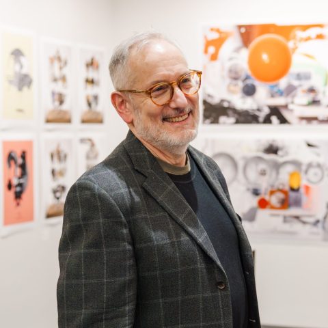 A man wearing glasses, a plaid blazer, and khaki pants stands smiling in an art gallery with contemporary artwork displayed on the walls behind him.