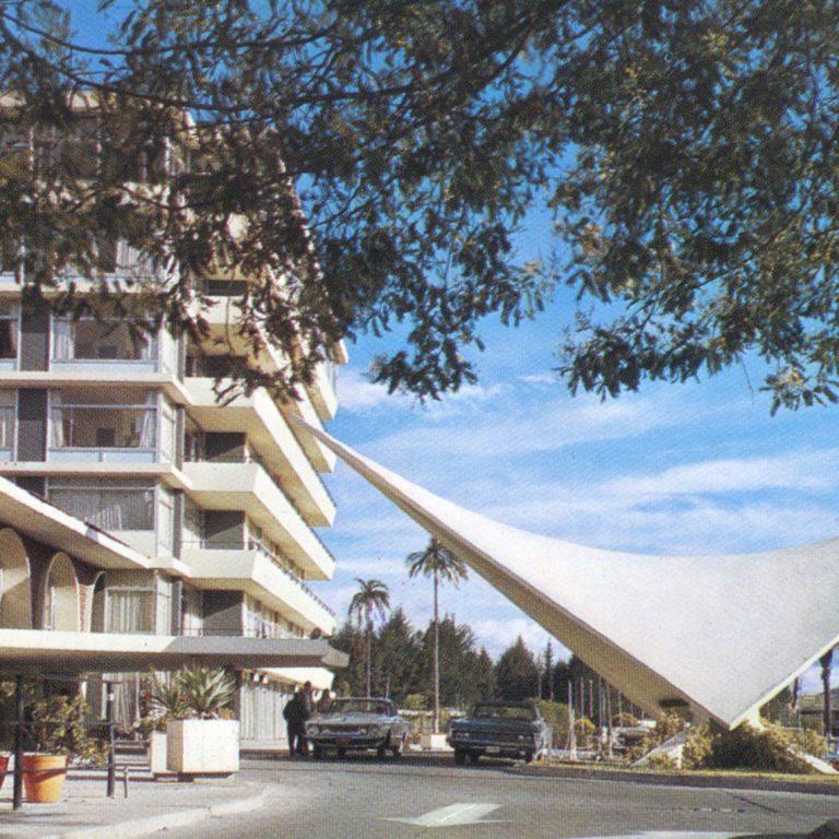 A mid-century modern hotel with a distinctive white, wing-shaped structure at the entrance, surrounded by palm trees and parked cars under a blue sky.
