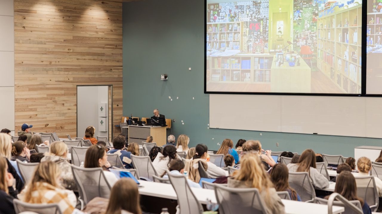 A university lecture hall with students seated, facing a lecturer at the front and a large screen displaying an image of a bookstore.
