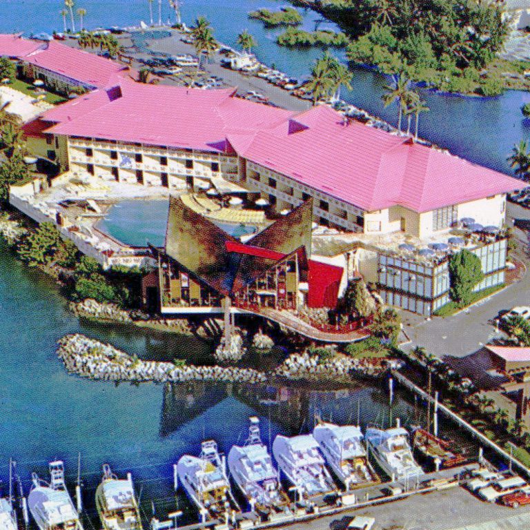Aerial view of a waterfront hotel with a pink roof, a central pool, boats docked nearby, and surrounding palm trees and water.