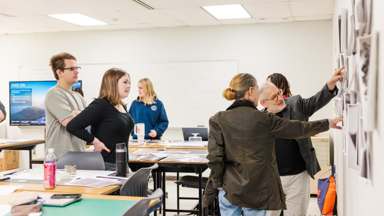 A group of people in a classroom discuss and examine photos displayed on a wall, with tables covered in papers and art supplies.