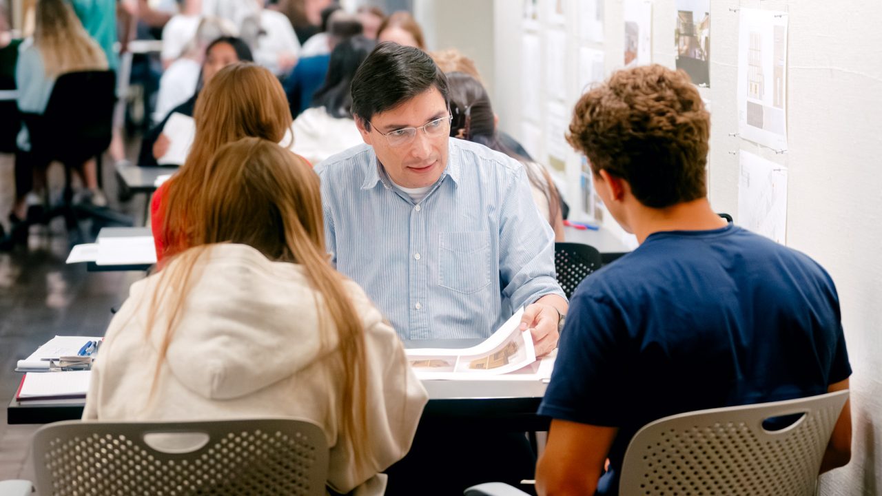 A man reviews documents with two students at a table in a busy hallway filled with people and architectural drawings on the walls.