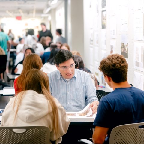 A man reviews documents with two students at a table in a busy hallway filled with people and architectural drawings on the walls.