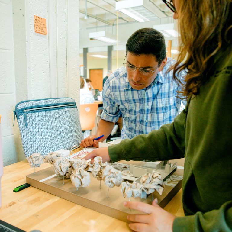 Two people examine a small architectural model with paper trees on a wooden table in a classroom or studio setting.