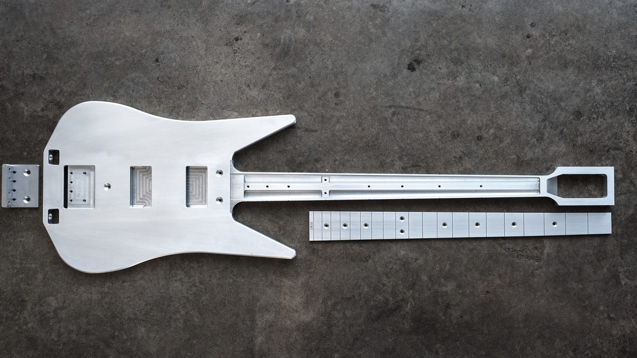 A metal electric guitar body and neck, unassembled, lying on a concrete surface with visible routing and drilled holes for hardware and components.
