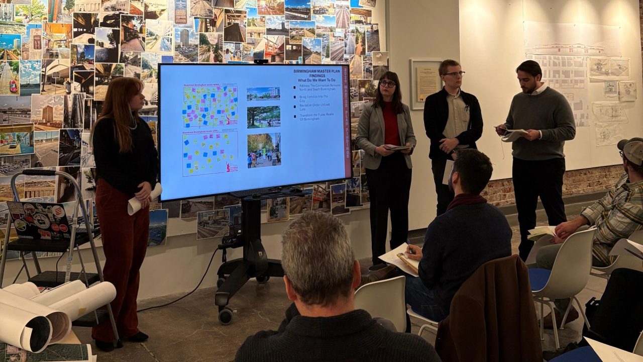 A group of four people stands beside a digital screen presenting to an audience in a modern, well-lit room with architectural designs displayed on the walls.