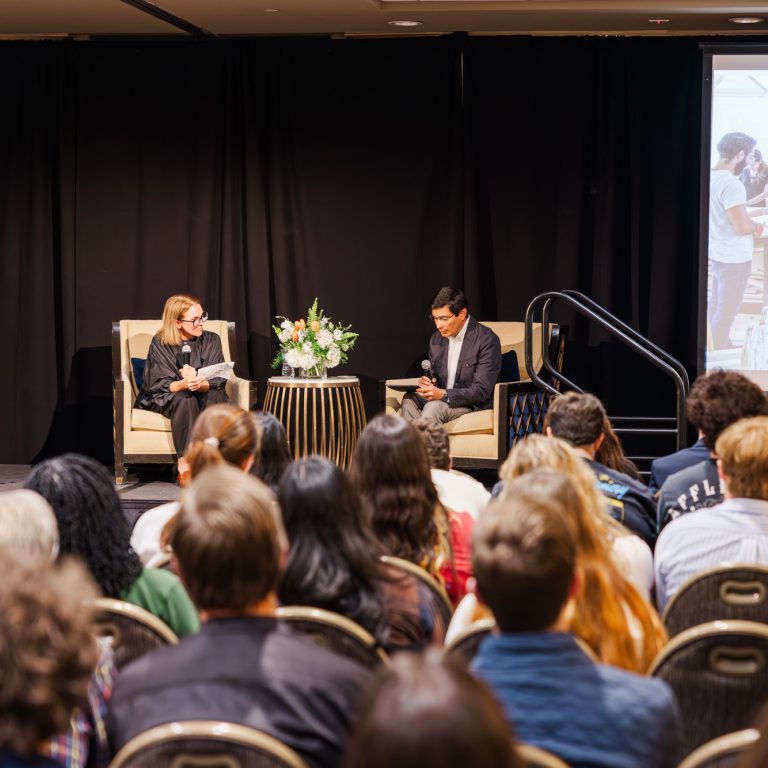Two people sit on stage having a discussion in front of an audience, with a podium, table with flowers, and a projected image visible in the background.