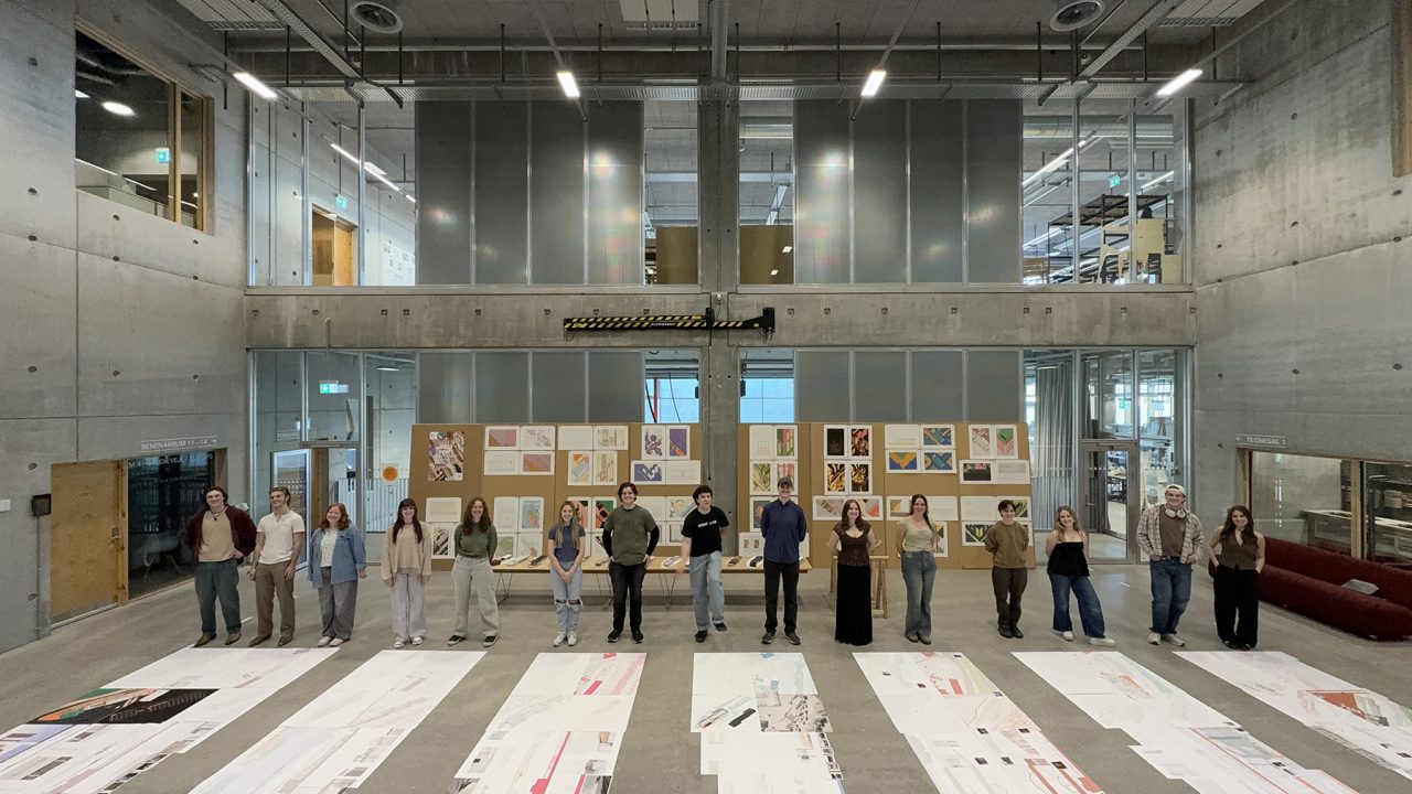 A group of people stands in a row inside a modern, industrial-style building, with large printed designs or plans laid out on the floor and posters displayed on the back wall.