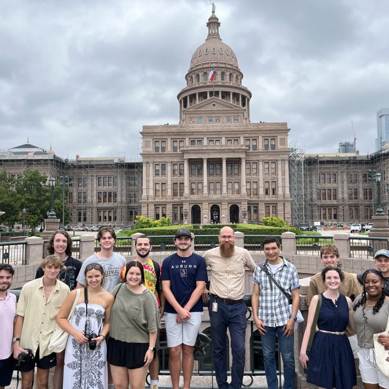 A group of people poses in front of the Texas State Capitol building on a cloudy day.