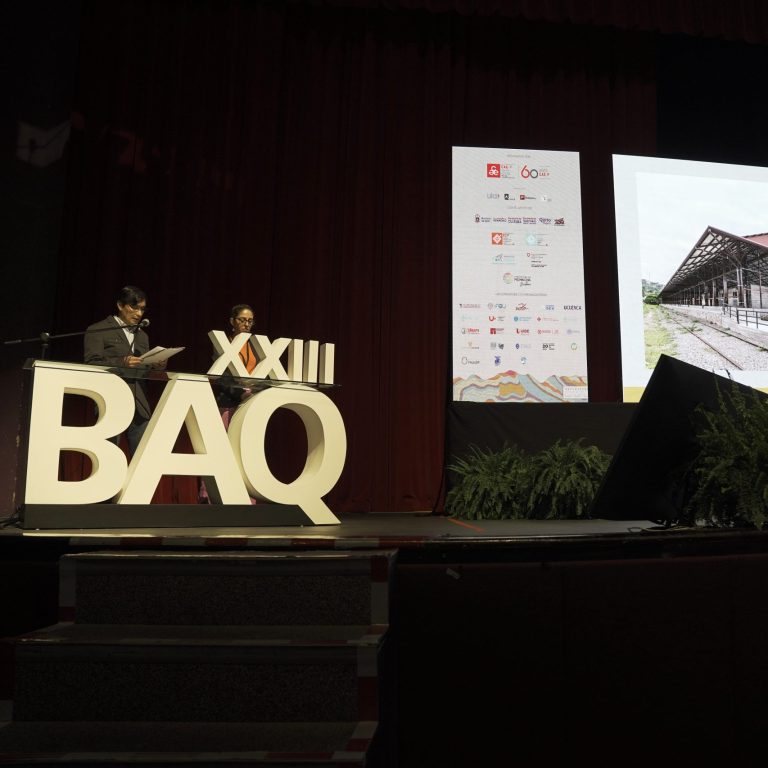 Two presenters stand at a podium onstage next to large XXIII BAQ letters, with a projected architectural image and text displayed on a screen in the background.