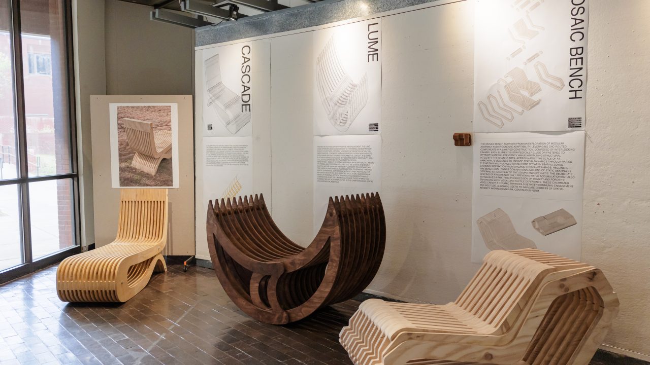 Three modern wooden benches with curved, layered designs are displayed in a gallery space, each with an explanatory panel mounted on the wall behind them.