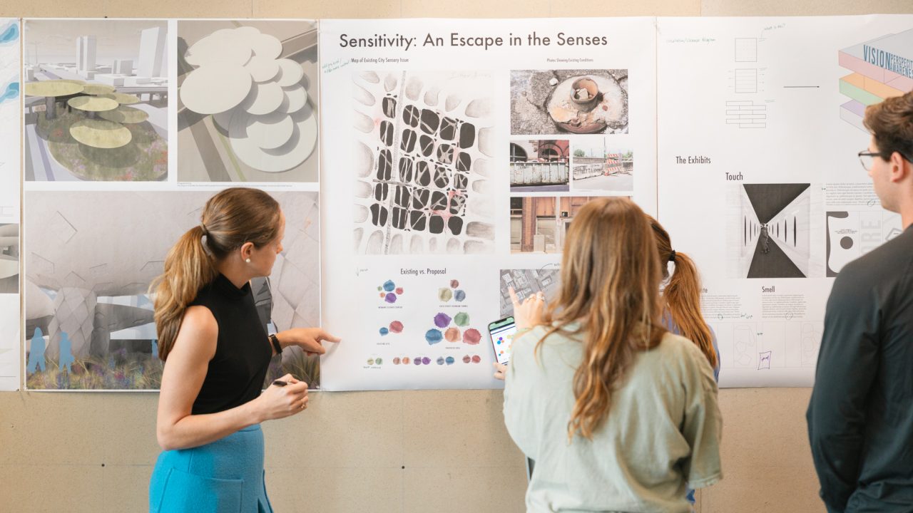 Four people stand in front of a wall displaying architectural design posters, discussing diagrams and images featured in a project titled “Sensitivity: An Escape in the Senses.”.