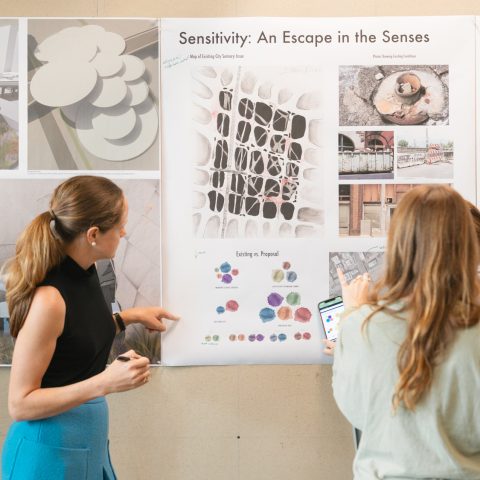 Four people stand in front of a wall displaying architectural design posters, discussing diagrams and images featured in a project titled “Sensitivity: An Escape in the Senses.”.