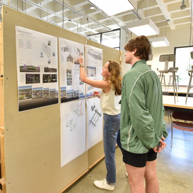 Two people stand in a classroom by a display board with architectural drawings and plans; one points at the drawings while the other observes.