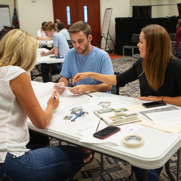Three people sit at a table covered with papers, tape, and a phone, engaged in discussion and working on a project in a classroom setting.