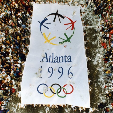 A large crowd surrounds a giant banner displaying the Atlanta 1996 Summer Olympics logo and rings, viewed from above.