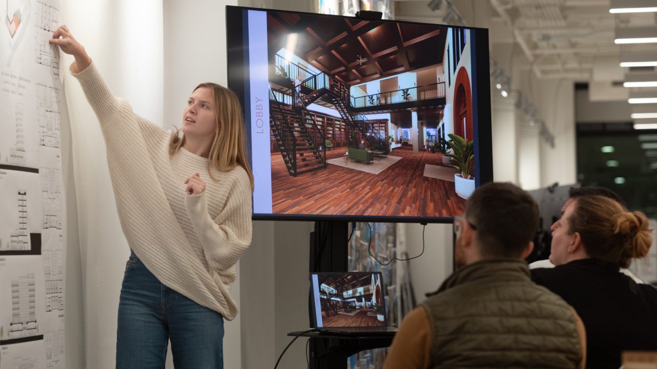A woman presents architectural plans and a digital rendering of a lobby to three seated people in a modern, well-lit office space.