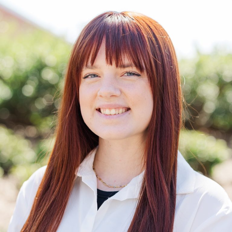 A young woman with long straight red hair and bangs, wearing a white button-up shirt, stands outdoors in front of greenery, smiling at the camera.