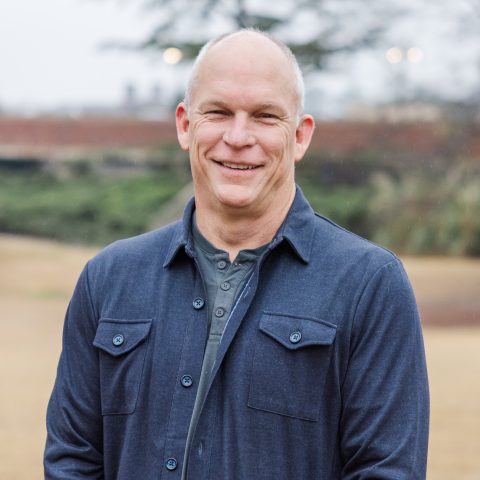 A middle-aged man with short hair smiles outdoors in a park-like setting on an overcast day. He is wearing a blue button-up shirt over a gray t-shirt.