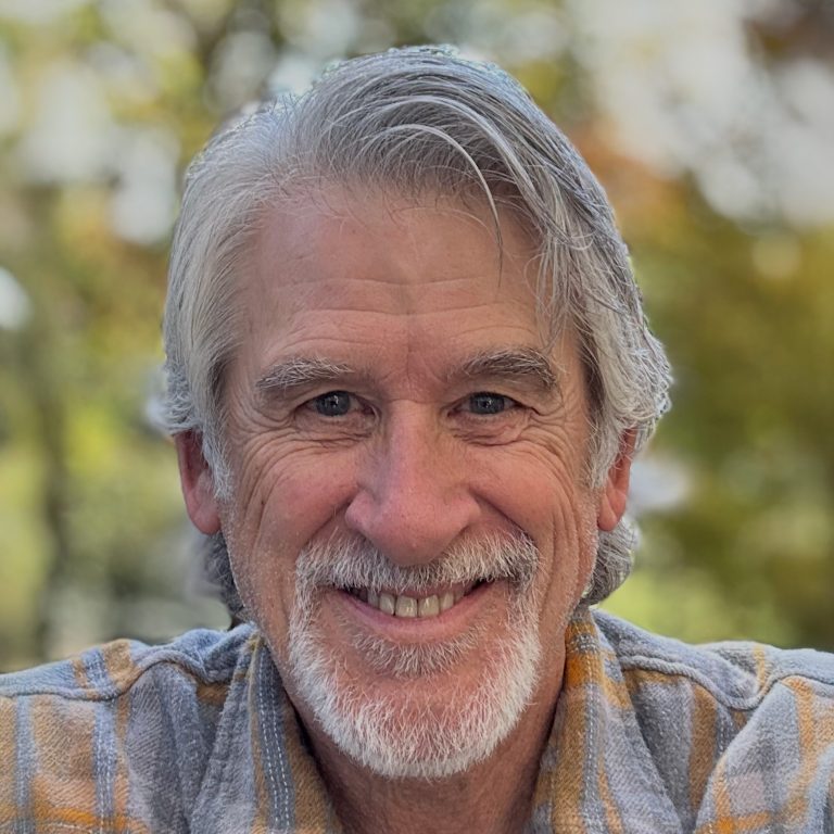An older man with gray hair and a beard smiles at the camera, wearing a plaid shirt outdoors with blurred greenery in the background.
