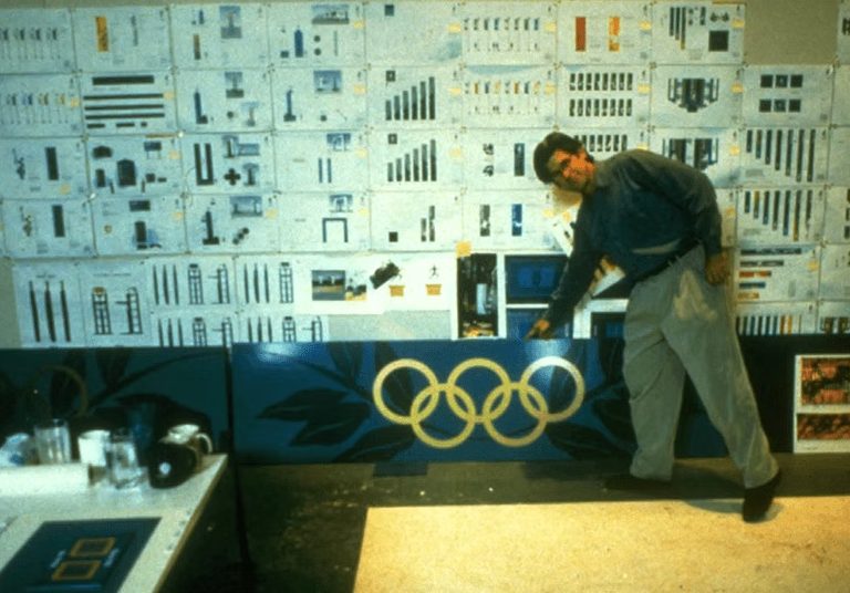 A person stands in front of a wall covered with design drafts, pointing at a panel featuring the Olympic rings.