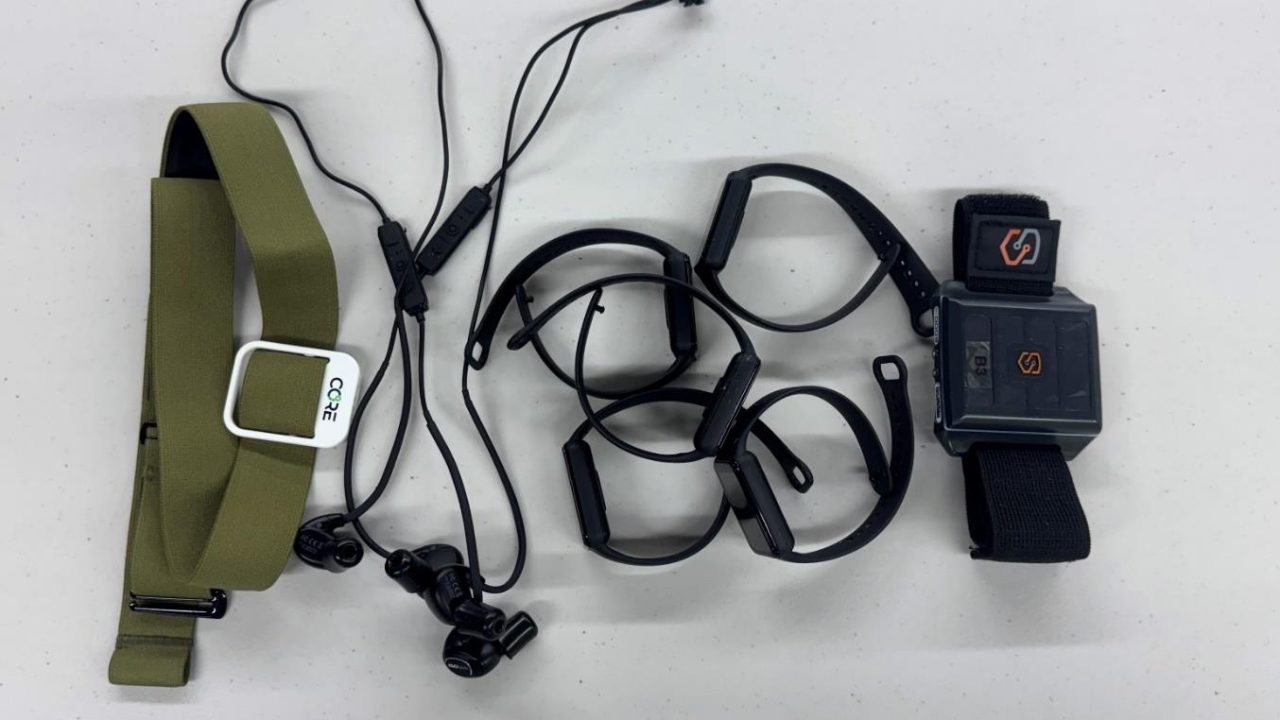 A green elastic headband, black wired earphones, black wristbands, and a black smartwatch with a logo are laid out on a white surface.