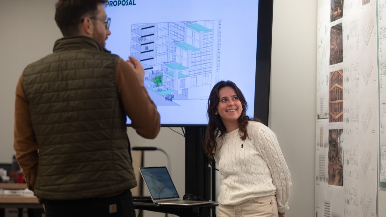Two people discuss an architectural proposal displayed on a screen in an office setting, with design plans visible on the wall.