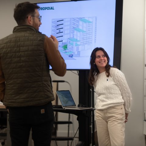 Two people discuss an architectural proposal displayed on a screen in an office setting, with design plans visible on the wall.