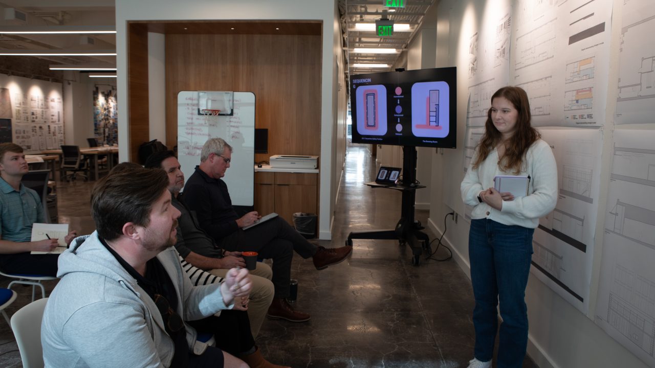 A woman stands by technical drawings and a presentation screen, speaking to a seated group in a modern office setting.
