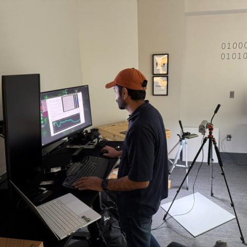 A man in a cap works at a standing desk with multiple monitors in an office; a tripod and scanning equipment are set up nearby.