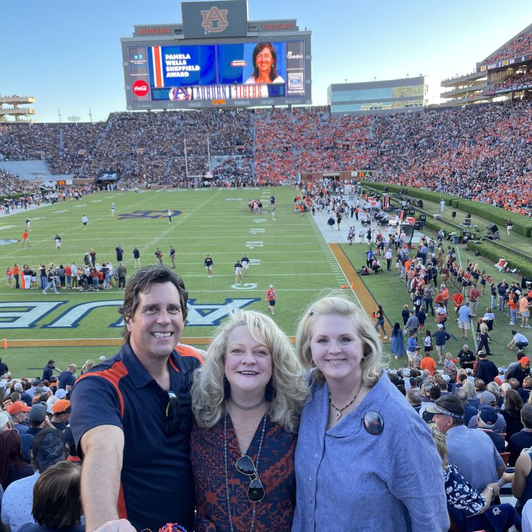 Three people smile for a photo in a stadium with a football game in progress; the stands are crowded and a scoreboard is visible in the background.