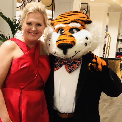 A woman in a red gown stands next to a person wearing a tiger mascot costume in a suit and bow tie, indoors on a hardwood floor.