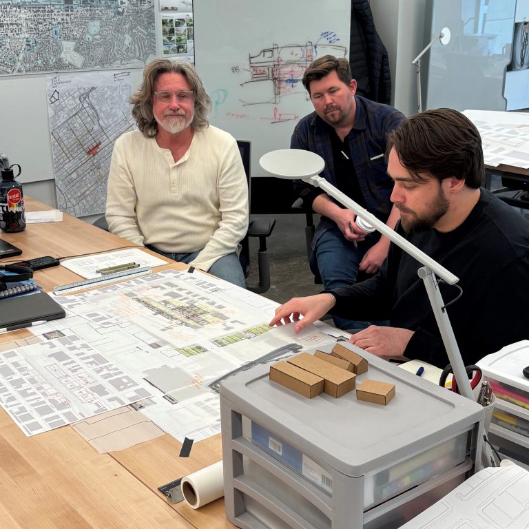 Three men review architectural plans and models at a workspace, surrounded by maps, sketches, and office supplies.