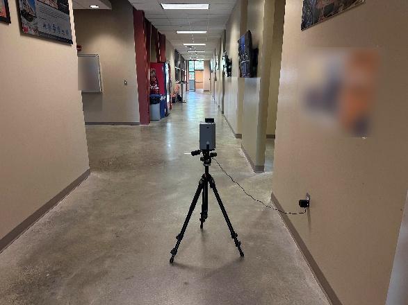 A smartphone mounted on a tripod is positioned in the middle of a hallway with beige walls and a polished floor. The phone is connected to a wall outlet with a cable.