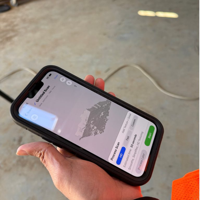 A hand holds a smartphone displaying a screen with a mountain image and location information, in a brightly lit indoor setting with a concrete floor.